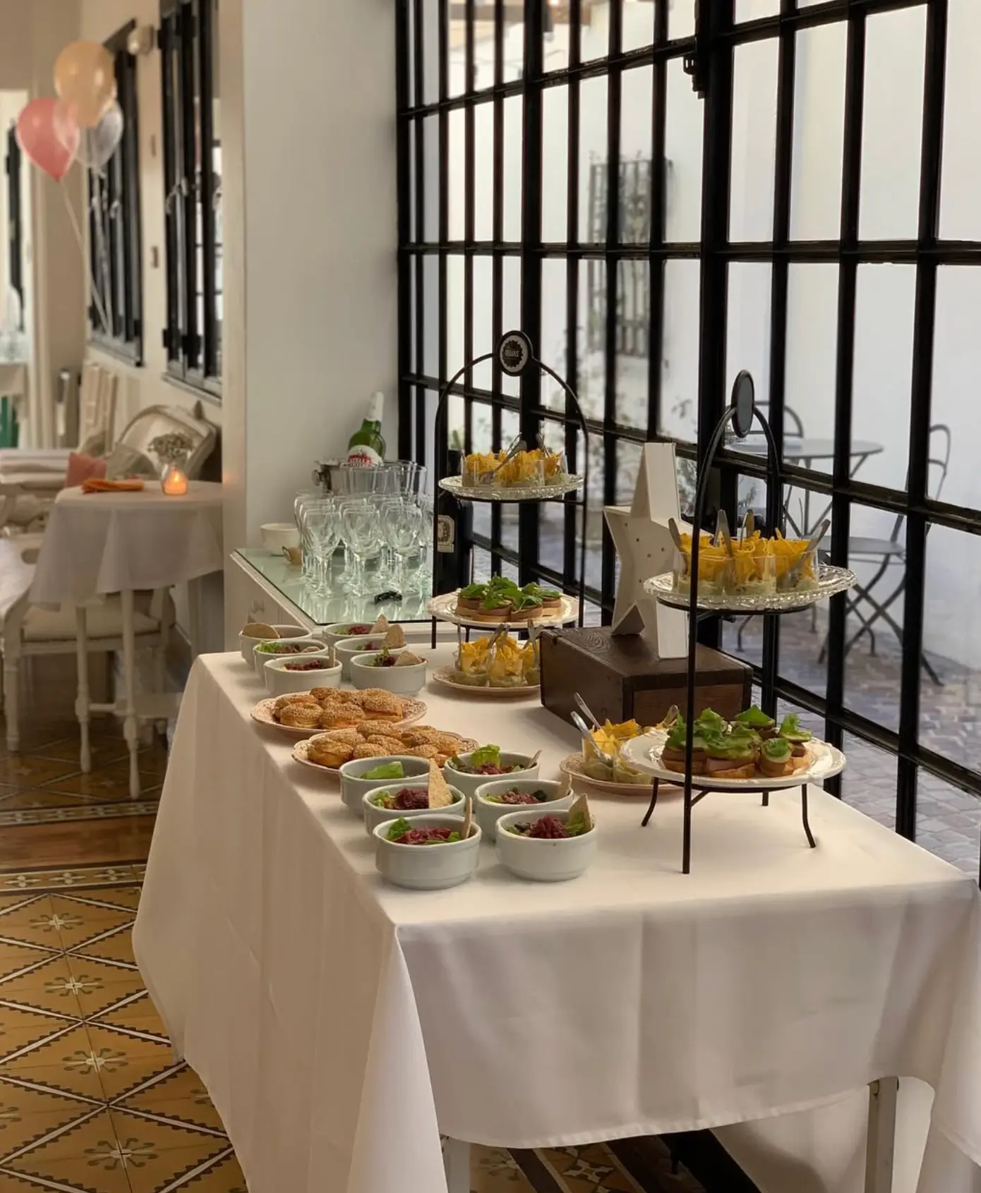 Mesa de recepción con bocados fríos y copas junto al ventanal de hierro